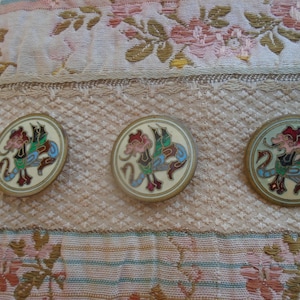 Peut inclure: Trois boutons ronds de style ancien avec un fond crème et un motif de dragon coloré. Le dragon présente des accents rouges, verts et bleus. Les boutons sont posés sur un tissu à motifs floraux.