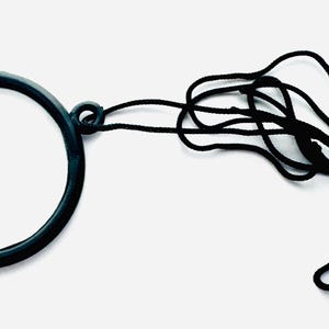 May include: A black monocle with a black cord attached. The monocle has a circular frame and a clear lens. The cord is knotted and looped. The background is white.