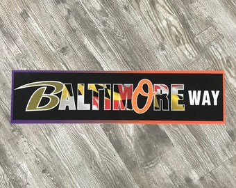May include: Black and orange banner with the text "Baltimore Way" in white letters. The "O" in Baltimore is replaced with the Maryland state flag.