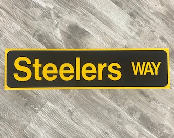 May include: A rectangular sign with a black background and a yellow border. The words "Steelers WAY" are printed in yellow, bold letters. The sign is likely a decorative item or a directional marker.