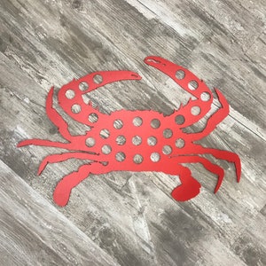 May include: A red metal crab wall art piece. The crab has a detailed design with cut-out circles throughout its body and claws. The crab is displayed on a wooden surface.