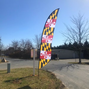May include: A tall, curved flag featuring the Maryland state flag design. The flag has a black and yellow checkered pattern with red and white crosses. It is displayed outdoors on a sunny day.