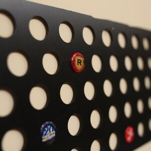 Peut inclure: Porte-capsule de bouteille en m&eacute;tal noir avec une grille de trous circulaires. Le porte-capsule est rempli de divers capsules de bouteille, dont une capsule rouge avec un logo "R" dor&eacute;.