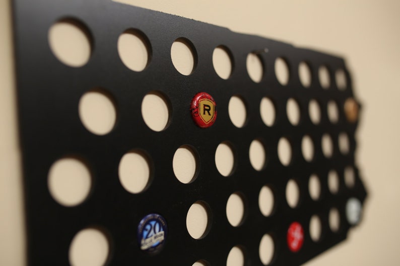 Peut inclure: Porte-capsule de bouteille en m&eacute;tal noir avec une grille de trous circulaires. Le porte-capsule est rempli de divers capsules de bouteille, dont une capsule rouge avec un logo "R" dor&eacute;.
