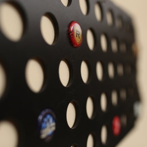 Peut inclure: Un porte-capsule en m&eacute;tal noir avec une grille de trous circulaires. Un bouchon de bouteille rouge avec un logo 'R' dor&eacute; est dans l'un des trous.