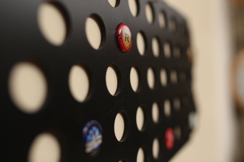 Peut inclure: Un porte-capsule en m&eacute;tal noir avec une grille de trous circulaires. Un bouchon de bouteille rouge avec un logo 'R' dor&eacute; est dans l'un des trous.