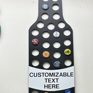 May include: Black bottle-shaped wall decor with circular openings to hold bottle caps. The sign has the words "CUSTOMIZABLE TEXT HERE" printed on it. Several colorful bottle caps are displayed in the openings.