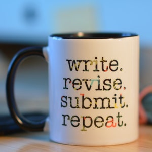 May include: White ceramic mug with a black rim and handle. The mug has the text "write. revise. submit. repeat." printed in black on the front.