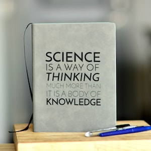 May include: A light gray notebook with the text "SCIENCE IS A WAY OF THINKING MUCH MORE THAN IT IS A BODY OF KNOWLEDGE" in black. A black ribbon bookmark and three pens, including a blue one, are visible.