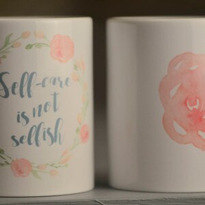 May include: Two white ceramic mugs with floral designs. One mug has a pink watercolor rose on the front. The other mug has a floral wreath with the text "Self-care is not selfish".