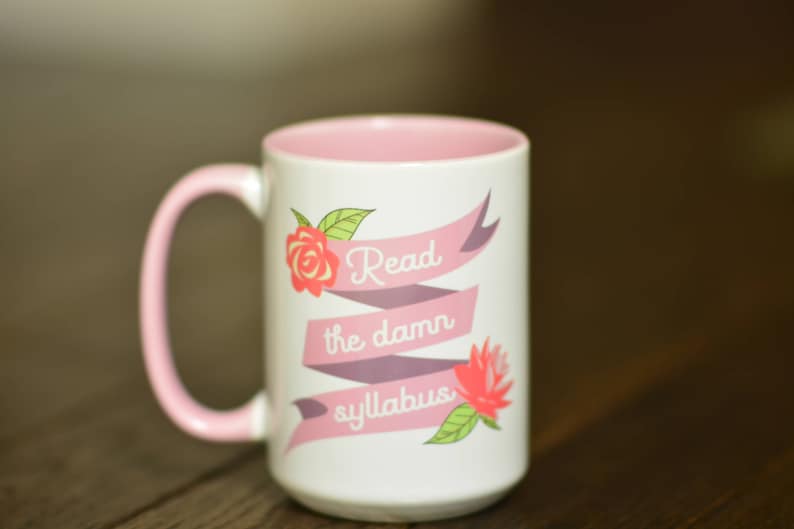 May include: A pink and white ceramic coffee mug with a pink rim and handle. The mug has a pink and purple ribbon with the text "Read the damn syllabus" and a pink rose and a pink flower with green leaves.