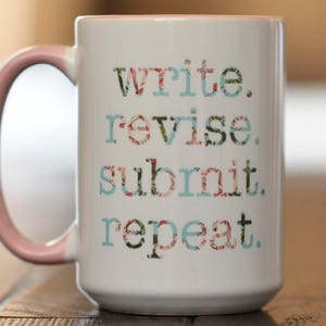 May include: A white ceramic mug with a pink handle. The mug has a floral design and the text "write. revise. submit. repeat." printed on it.