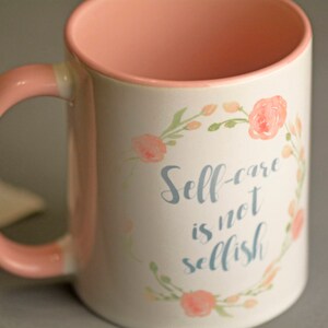 May include: A pink and white ceramic mug with a floral design and the text "Self-care is not selfish".