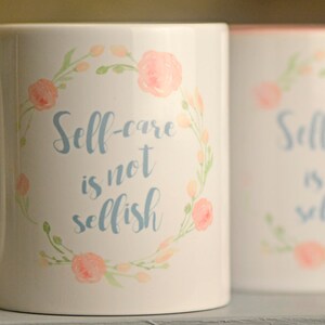 May include: Two white ceramic mugs with a floral design and the text "Self-care is not selfish" in blue lettering.