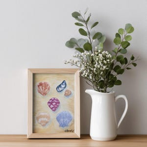 May include: A framed watercolor painting of various seashells in shades of orange, blue, and purple. The painting is in a light wooden frame and is displayed on a wooden surface. A white pitcher with green foliage and white flowers is to the right.