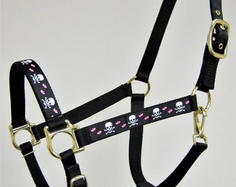 May include: Black horse halter with a pink and white skull and heart design. The halter has a gold buckle and rings.