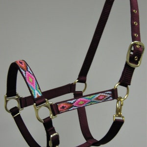 Puede incluir: Un cabezal de caballo burdeos con una correa estampada en rosa, azul y blanco. El cabezal tiene herrajes de metal dorado, incluyendo una hebilla y anillos.