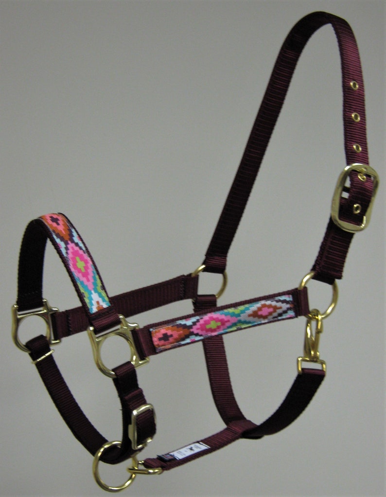 Puede incluir: Un cabezal de caballo burdeos con una correa estampada en rosa, azul y blanco. El cabezal tiene herrajes de metal dorado, incluyendo una hebilla y anillos.