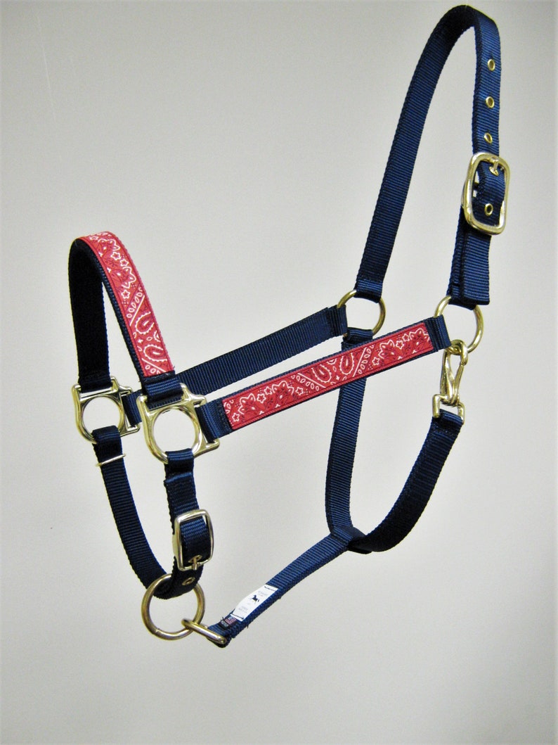 Puede incluir: Un cabezal para caballo azul marino con estampado de bandana rojo. El cabezal tiene una hebilla de lat&oacute;n y dos anillos de lat&oacute;n.