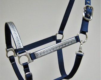 May include: A navy blue horse halter with silver accents. The halter has a buckle on the side and a metal ring on the nose.
