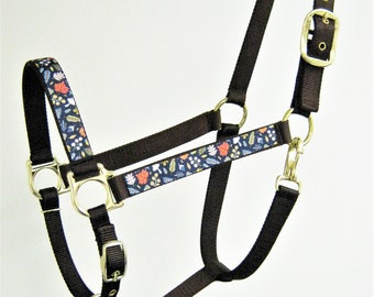 May include: A brown nylon horse halter with a floral print on the noseband and cheek pieces. The halter has gold-colored hardware, including a buckle, rings, and a snap hook.