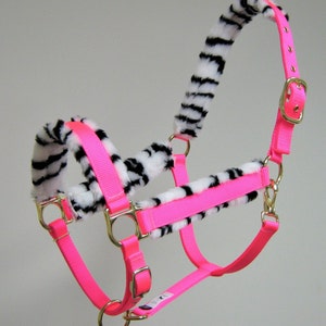 May include: A pink horse halter with a zebra print fleece lining. The halter has a buckle on the side and a metal ring on the noseband.