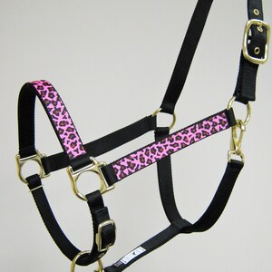 Pode incluir: Um cabresto de cavalo de nylon preto com uma guarni&ccedil;&atilde;o de estampa de leopardo rosa e marrom. O cabresto tem uma fivela e an&eacute;is dourados.
