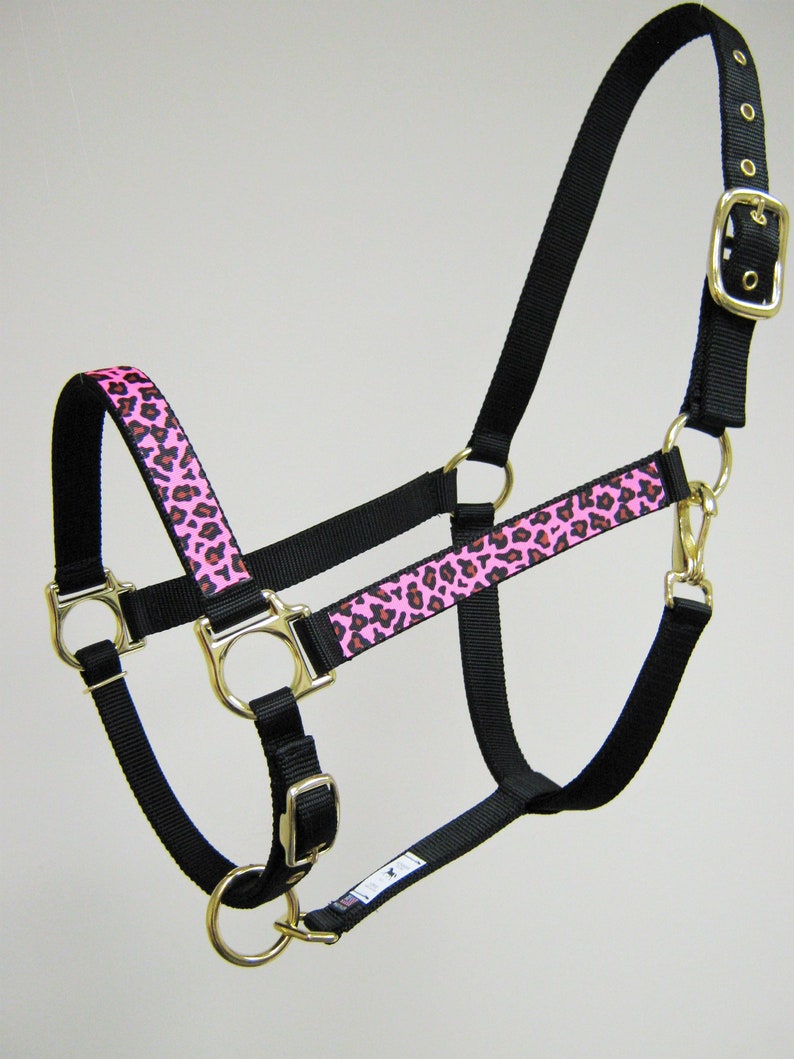 Pode incluir: Um cabresto de cavalo de nylon preto com uma guarni&ccedil;&atilde;o de estampa de leopardo rosa e marrom. O cabresto tem uma fivela e an&eacute;is dourados.