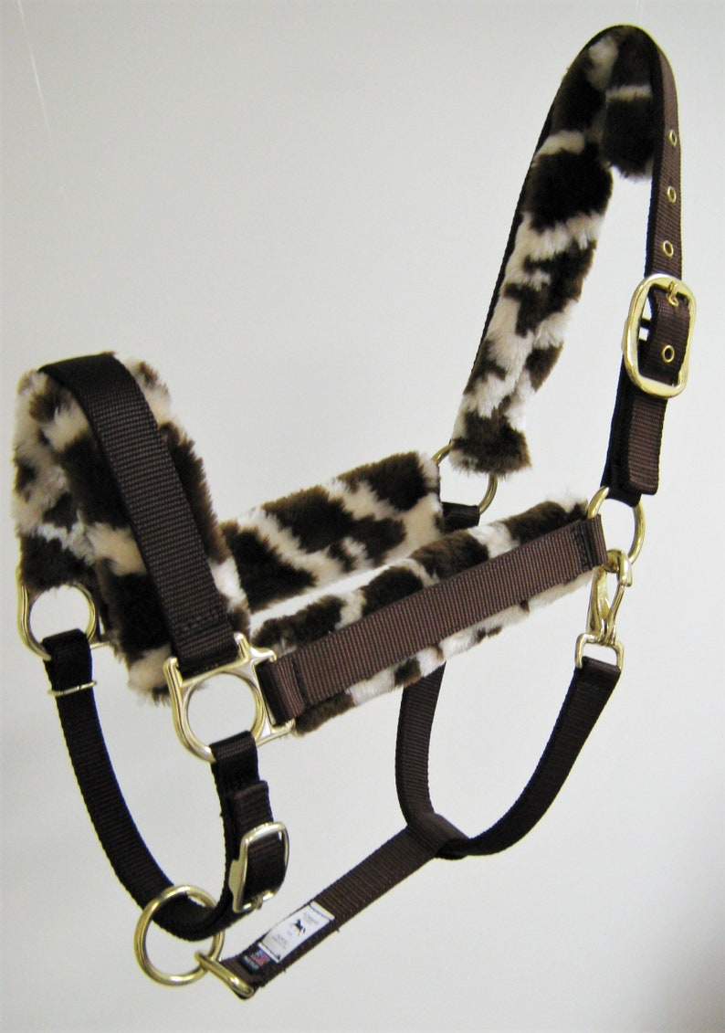 May include: Brown and white horse halter with fleece lining. The halter has a buckle on the side and a snap on the chin.