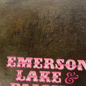 May include: Close-up of a vintage album cover with the band name "Emerson Lake & Palmer" in pink, ornate lettering. The cover has signatures in black ink. The background is a textured, dark brown color.