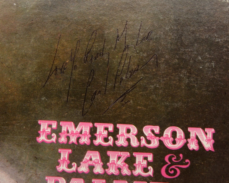 May include: Close-up of a vintage album cover with the band name "Emerson Lake & Palmer" in pink, ornate lettering. The cover has signatures in black ink. The background is a textured, dark brown color.