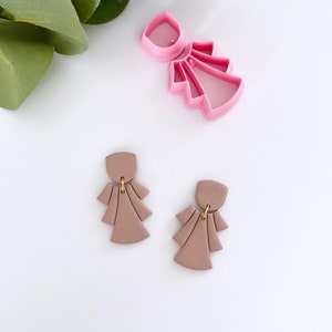 May include: A pair of mauve polymer clay earrings with a geometric fan shape and gold jump rings. The earrings are shown next to a pink plastic cutter.