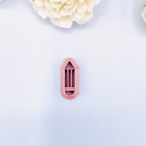 May include: A pink plastic cookie cutter shaped like a pencil. The cutter has a detailed design with a pointed tip and a flat base.