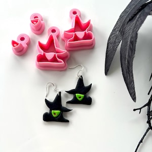 May include: Pink witch hat-shaped cookie cutters and black witch hat earrings with green accents. The earrings have silver hooks. The cookie cutters are in various sizes. A black feather and branch are in the background.