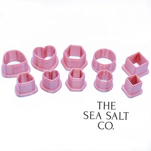May include: A set of ten pink plastic cookie cutters in various shapes, including hearts, circles, squares, and hexagons. The cutters are arranged on a white surface. The text "THE SEA SALT CO." is printed below the cutters.