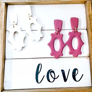 May include: Two pairs of statement earrings, one pair is white and the other is a deep burgundy red. The earrings are displayed on a white wooden surface with the word "love" written in black.