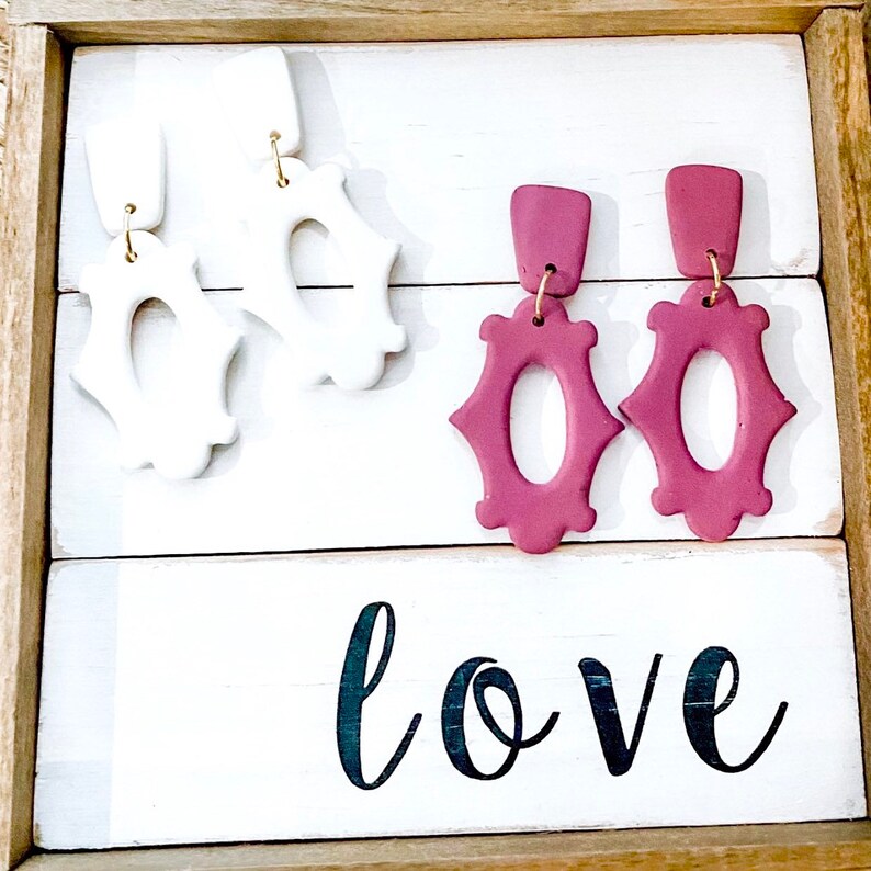 May include: Two pairs of statement earrings, one pair is white and the other is a deep burgundy red. The earrings are displayed on a white wooden surface with the word "love" written in black.