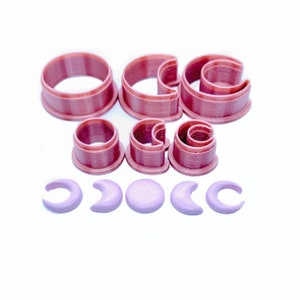 May include: A set of pink plastic cookie cutters in the shape of crescent moons and circles. The cutters are arranged in a pattern on a white surface.