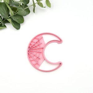 May include: Pink plastic cookie cutter shaped like a crescent moon with a spiderweb design.