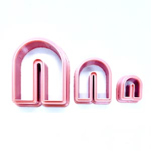 May include: Three pink plastic cookie cutters shaped like arches, in descending sizes.