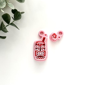 May include: Two pink plastic cookie cutters, one shaped like a tiki head with a straw and the other shaped like a flower.