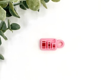 May include: A pink plastic charm with a small heart and three rectangular cutouts. The charm has a loop for attaching to a keychain or other items.