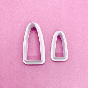 May include: Two white plastic cookie cutters shaped like arches. The larger arch is about twice the size of the smaller arch.
