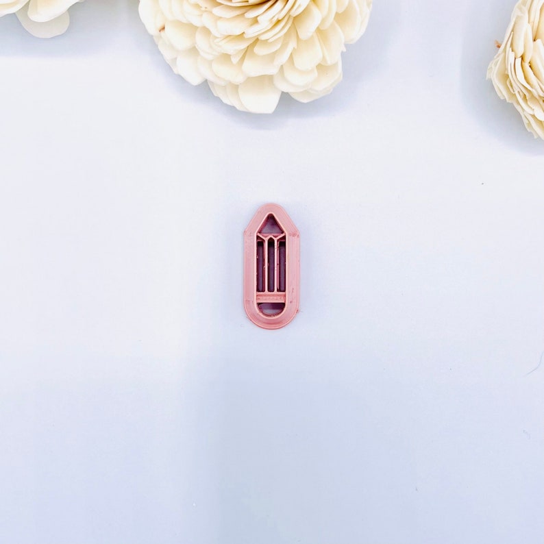 May include: A pink plastic cookie cutter shaped like a pencil. The cutter has a detailed design with a pointed tip and a flat base.