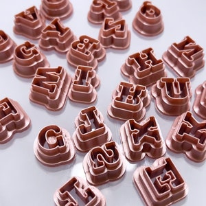 May include: A selection of rose-gold letter-shaped cookie cutters on a white plate. The cutters are in capital letter shapes, suitable for baking or crafts. The image is well-lit, showing the metallic finish of the cutters.