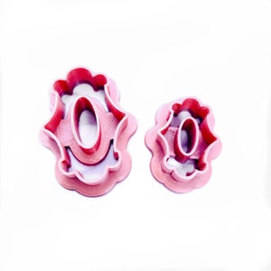 May include: Two pink plastic cookie cutters shaped like the letter "O" with a decorative design. The larger cutter is slightly larger than the smaller cutter.