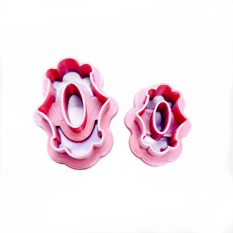 May include: Two pink plastic cookie cutters shaped like the letter "O" with a decorative design. The larger cutter is slightly larger than the smaller cutter.