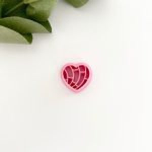 May include: Pink plastic cookie cutter shaped like a heart with a detailed design of a concha, a traditional Mexican sweet bread.