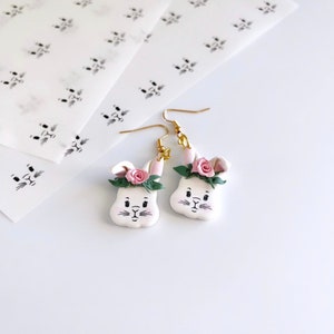 May include: A pair of white bunny-shaped earrings with pink flowers and green leaves. The earrings have gold hooks.