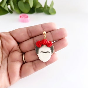 May include: A pair of gold stud earrings featuring a white portrait of Frida Kahlo with a red flower crown.